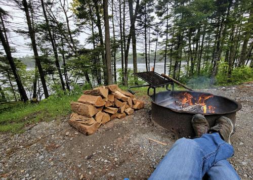 A person relaxes by a campfire near a stack of firewood, surrounded by trees and a lake in the background.