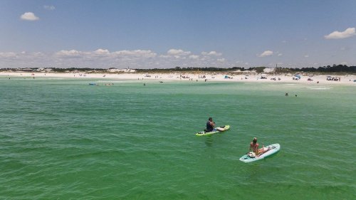 Two people paddleboarding on a clear green ocean, with a sandy beach and clouds in the background.