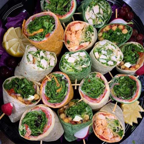 A colorful platter of assorted wraps filled with greens, chicken, and other ingredients, garnished with lemon and fruit.