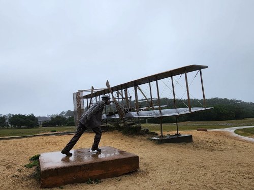 A statue of a man pushing a vintage airplane on a rainy day, set in a grassy area.