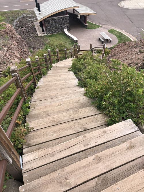 Wooden stairs descend a hillside, surrounded by greenery, leading to a pathway and a small shelter in the background.