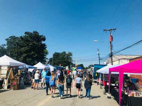 A bustling outdoor market scene with people walking between vendor tents under a clear blue sky.