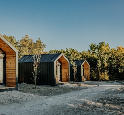Three modern cabins with wooden and black exteriors, set in a natural landscape with trees and a clear blue sky.