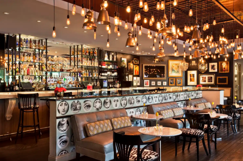 A stylish bar interior with a well-lit atmosphere, featuring a long counter, seating, and artistic decor on the walls.