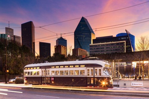 A vintage streetcar travels along a city street at sunset, with modern skyscrapers in the background.