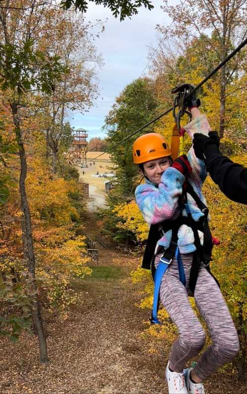 A girl in a colorful sweater and helmet zip lines through a forest with autumn foliage, smiling at the camera.