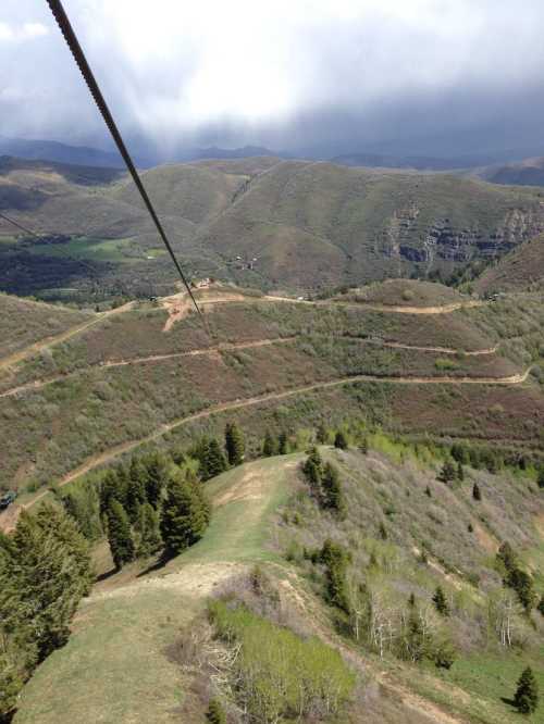 Aerial view of rolling green hills and valleys under a cloudy sky, with a zip line cable stretching across the scene.