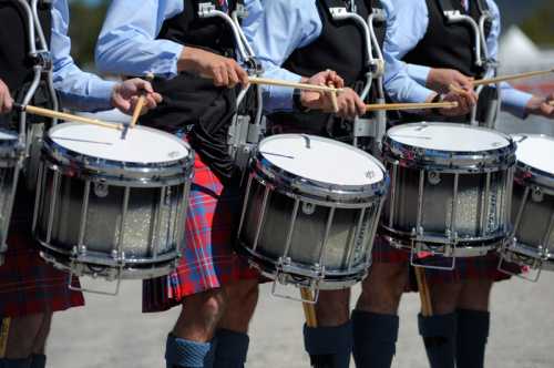 A line of drummers in kilts playing snare drums, holding drumsticks, with a clear blue sky in the background.