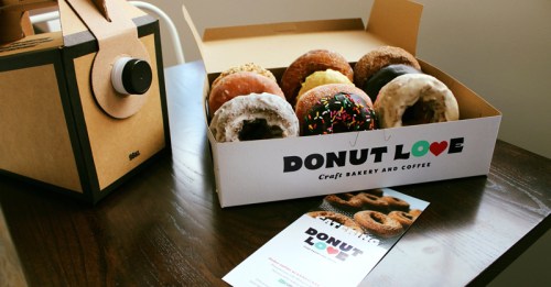 A box of assorted donuts from Donut Love, with a coffee box and promotional flyer on a wooden table.