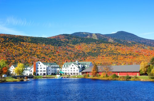 A scenic view of a lake with a white hotel and red barn, surrounded by vibrant autumn foliage and mountains.