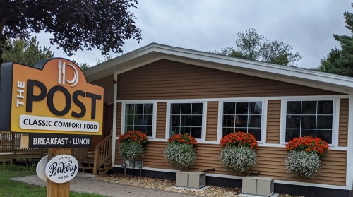 A cozy restaurant exterior with a sign reading "The Post" and flower planters, promoting breakfast and lunch.