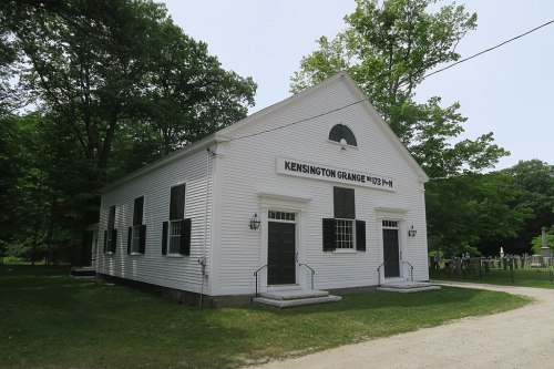 A white, historic building with black shutters and a sign reading "Kensington Grange M173 P.H." surrounded by trees.