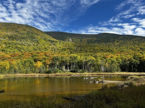 A serene landscape featuring a calm pond surrounded by vibrant autumn foliage and mountains under a blue sky.