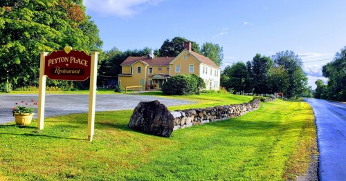 A yellow restaurant building with a sign, surrounded by greenery and a stone wall, beside a quiet road.