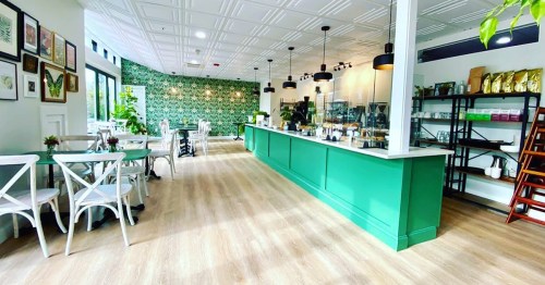 Bright café interior with green accents, wooden floors, and plants, featuring tables, chairs, and a service counter.