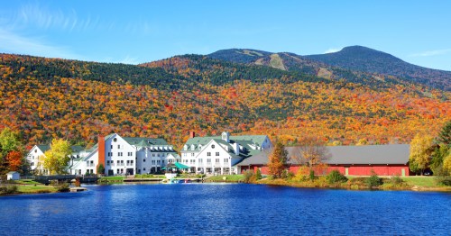 A scenic view of a lakeside hotel surrounded by vibrant autumn foliage and mountains under a clear blue sky.