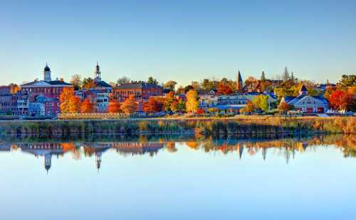 A serene lakeside view of a colorful autumn town, reflecting buildings and trees in calm water under a clear sky.