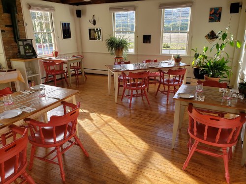 Bright restaurant interior with wooden floors, tables set with red chairs, and large windows letting in natural light.