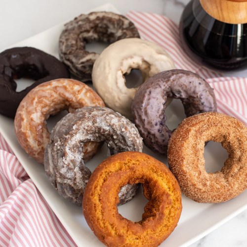 A variety of colorful donuts arranged on a plate, with a coffee pot in the background and a striped napkin.