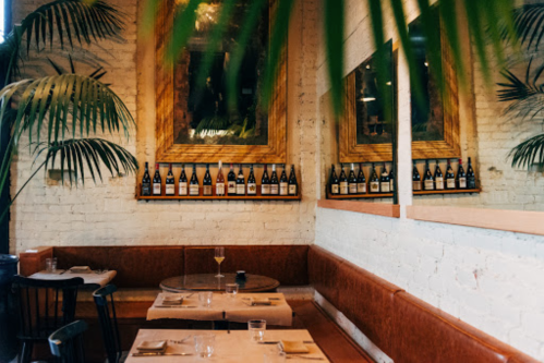 Cozy restaurant interior with green plants, wooden decor, and a wall of wine bottles above a brown leather booth.