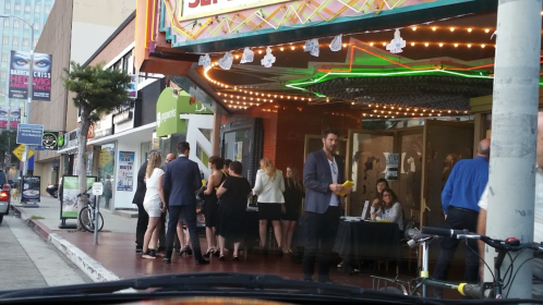 A group of people in formal attire gathers outside a theater, with bright lights and a festive atmosphere.
