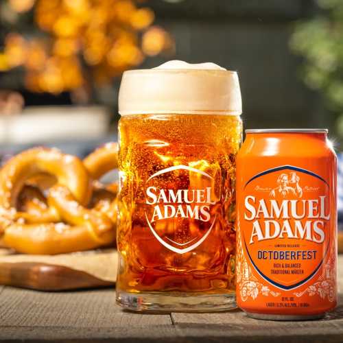 A glass of Samuel Adams OctoberFest beer next to a can and pretzels, with autumn foliage in the background.