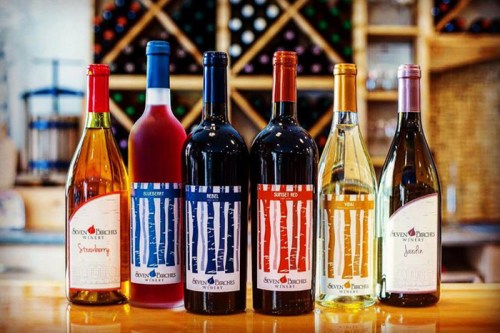 A display of six wine bottles in various colors, labeled with names, set against a wooden backdrop.