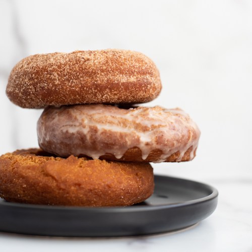 Three assorted donuts stacked on a black plate, featuring a glazed donut, a cinnamon sugar donut, and a pumpkin donut.