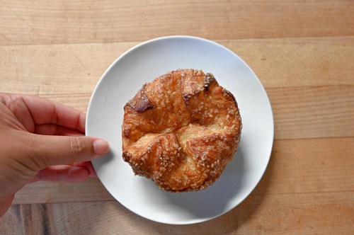 A hand holding a plate with a golden-brown pastry topped with sugar, resting on a wooden surface.