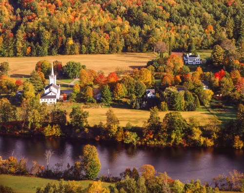 A scenic view of a river surrounded by vibrant autumn foliage, featuring a church and houses in a rural landscape.