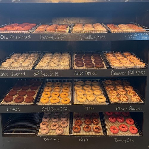 A display case filled with various donuts, including glazed, frosted, and specialty flavors, labeled with prices.