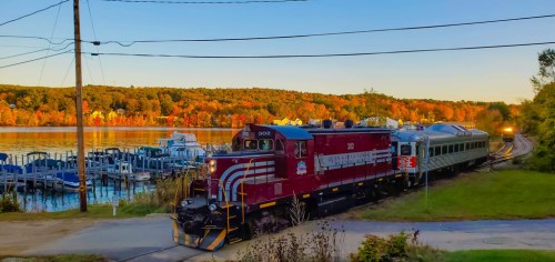 A red train with passenger cars travels along a scenic lakeside route, surrounded by autumn foliage.