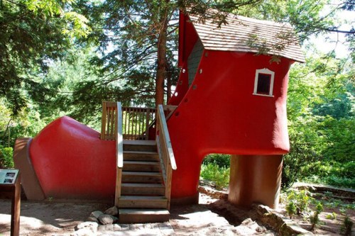 A whimsical red shoe-shaped house with a wooden staircase, surrounded by lush greenery and trees.