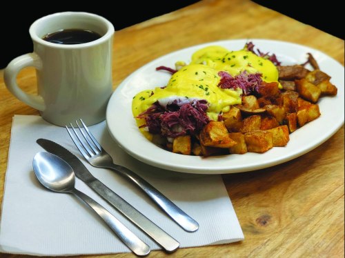 A plate of eggs Benedict with pulled meat and potatoes, accompanied by a cup of coffee and cutlery on a napkin.