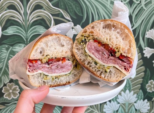 A hand holding a plate with a halved sandwich filled with meats, cheese, and colorful vegetables against a leafy background.