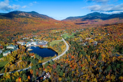 Aerial view of a vibrant autumn landscape with colorful trees, a pond, and a winding road surrounded by mountains.