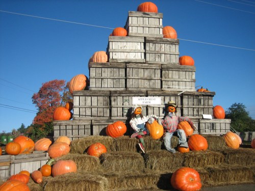 A pyramid of wooden crates stacked with pumpkins, surrounded by hay bales and two scarecrows sitting in front.