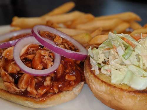 A close-up of a BBQ chicken sandwich with red onion slices, served with coleslaw and French fries on the side.