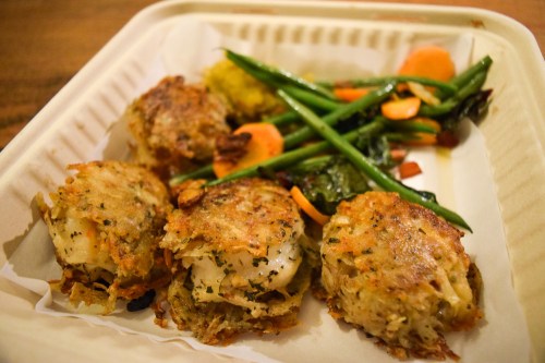 A takeout container with four golden-brown crab cakes and a side of sautéed vegetables, including green beans and carrots.