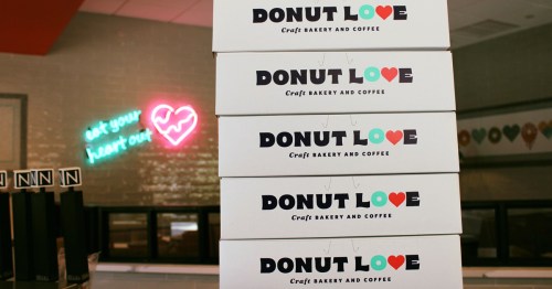Stack of white donut boxes labeled "DONUT LOVE" in a bakery with a neon heart sign in the background.