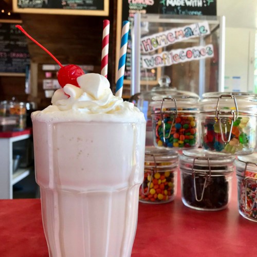 A creamy milkshake topped with whipped cream and a cherry, with colorful candies in jars in the background.