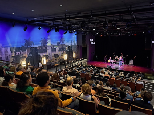 Audience seated in a theater with a stage set for a performance, featuring a colorful backdrop and band setup.