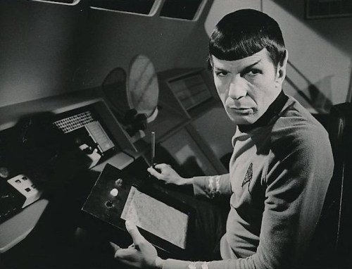 A black and white image of a man in a Starfleet uniform, holding a device, seated at a futuristic control panel.