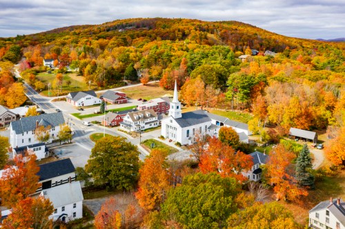 Aerial view of a quaint village surrounded by vibrant autumn foliage and a church with a tall steeple.