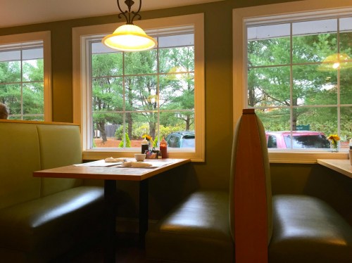 Cozy restaurant interior with green booths, tables, and large windows showing trees and parked cars outside.
