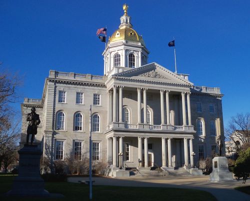 The New Hampshire State House, featuring a golden dome and columns, with a statue in the foreground against a clear blue sky.
