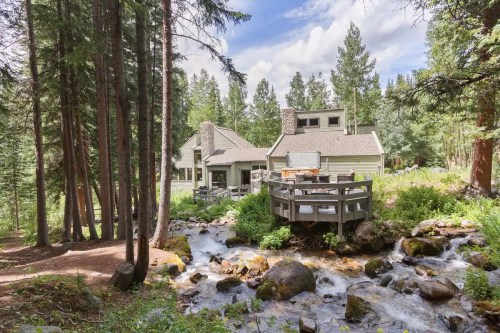 A cozy cabin nestled among trees, with a deck overlooking a flowing stream and rocky landscape.