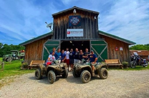 A group of people poses in front of a rustic barn with ATVs parked outside on a sunny day.