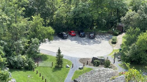 Aerial view of a parking lot surrounded by trees, with several parked cars and a pathway leading into the greenery.