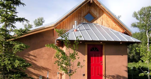 A small, rustic house with a red door, wooden accents, and a metal roof, surrounded by greenery.